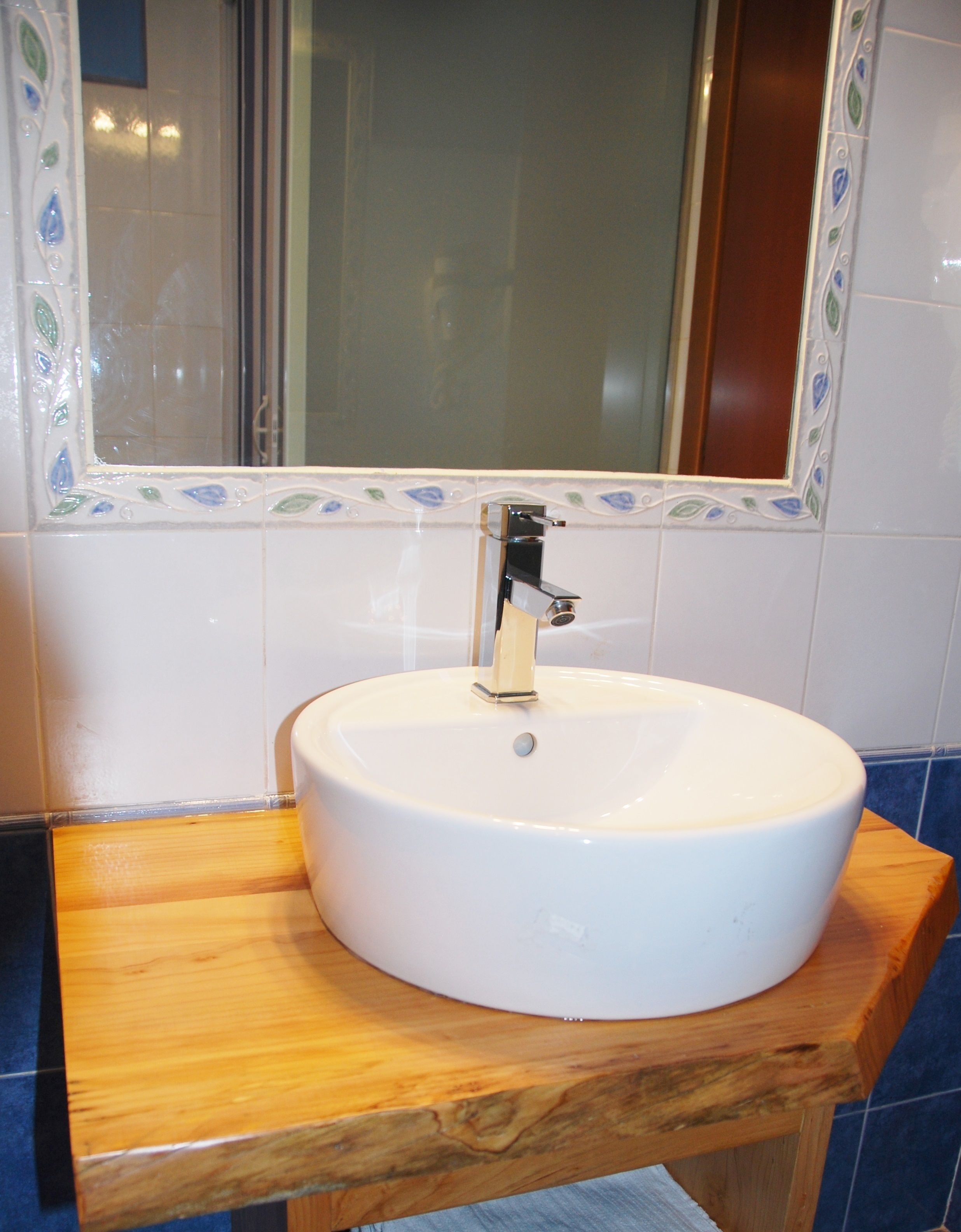 Ein Badezimmer mit einem weißen Waschbecken auf einer Holzausstattung, blauen Fliesen und einem Spiegel mit dekorativen Fliesen.
