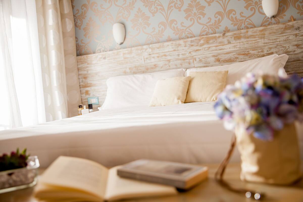 A neatly made bed with white linens and two pillows, a bedside table with a book and a vase of flowers, and a wall with a decorative pattern and a wall sconce.
