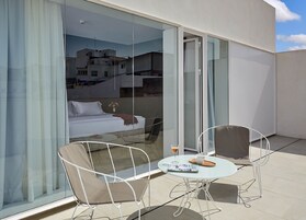 Un balcón moderno al aire libre con un ventanal de vidrio que da a un dormitorio. Hay dos sillas blancas y una pequeña mesa redonda con superficie de vidrio.