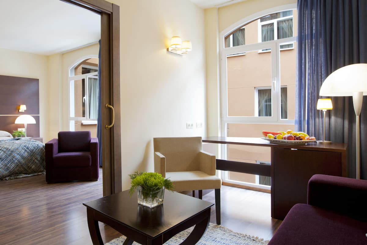 A hotel room with a bed, two armchairs, a small table with a vase, a balcony with a fruit bowl, and a view of the building outside.