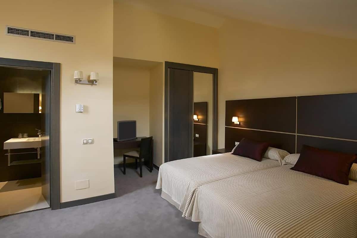 A hotel room with a bed, a desk, a chair, a mirror, and a bathroom with a sink.