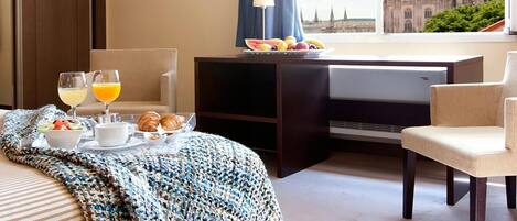 A hotel room with a bed, a desk with a computer, a chair, a view of a cathedral, and breakfast items on the bed.