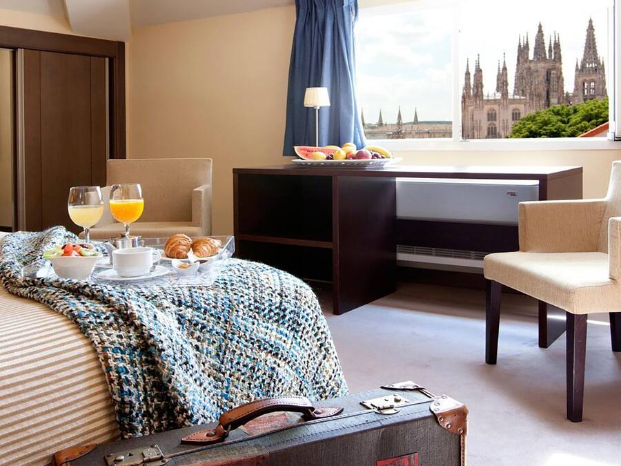Een hotelkamer met een bed, een bureau met een computer, een stoel, uitzicht op een kathedraal en ontbijtartikelen op het bed.