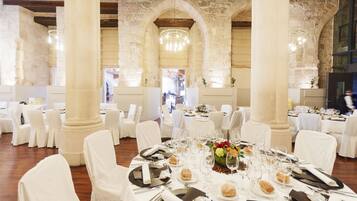 Un salón de banquetes con mesas redondas dispuestas para un evento formal, con sillas blancas y un arco de piedra en el fondo.