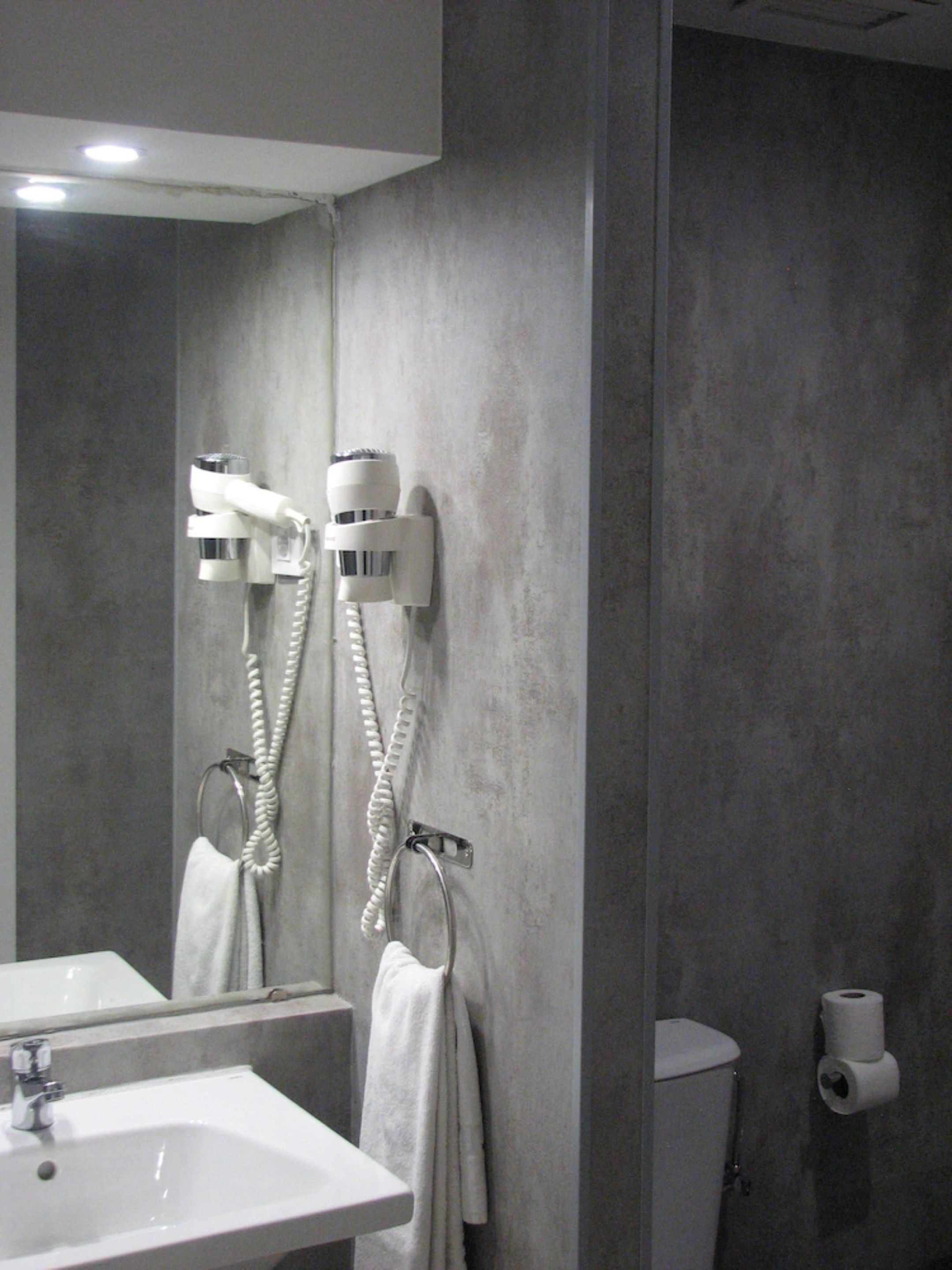 Un bagno con lavandino, specchio, portasciuga, asciugacapelli e dispenser di carta igienica.