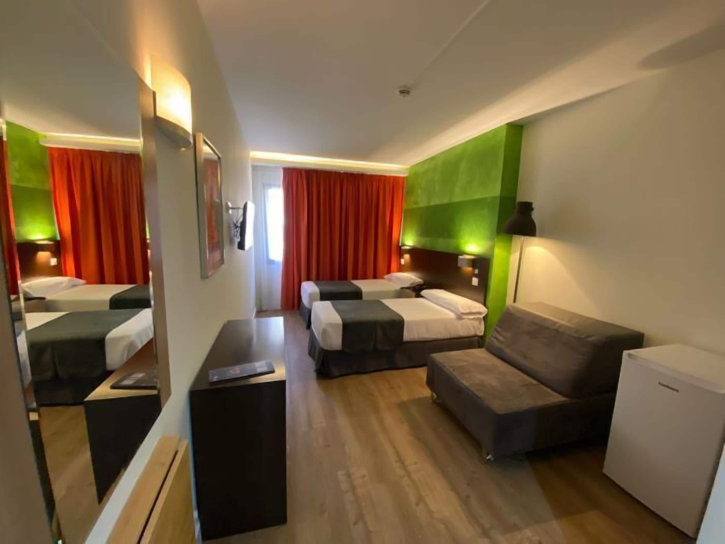 A hotel room with two beds, a sofa, a desk, and a small refrigerator.