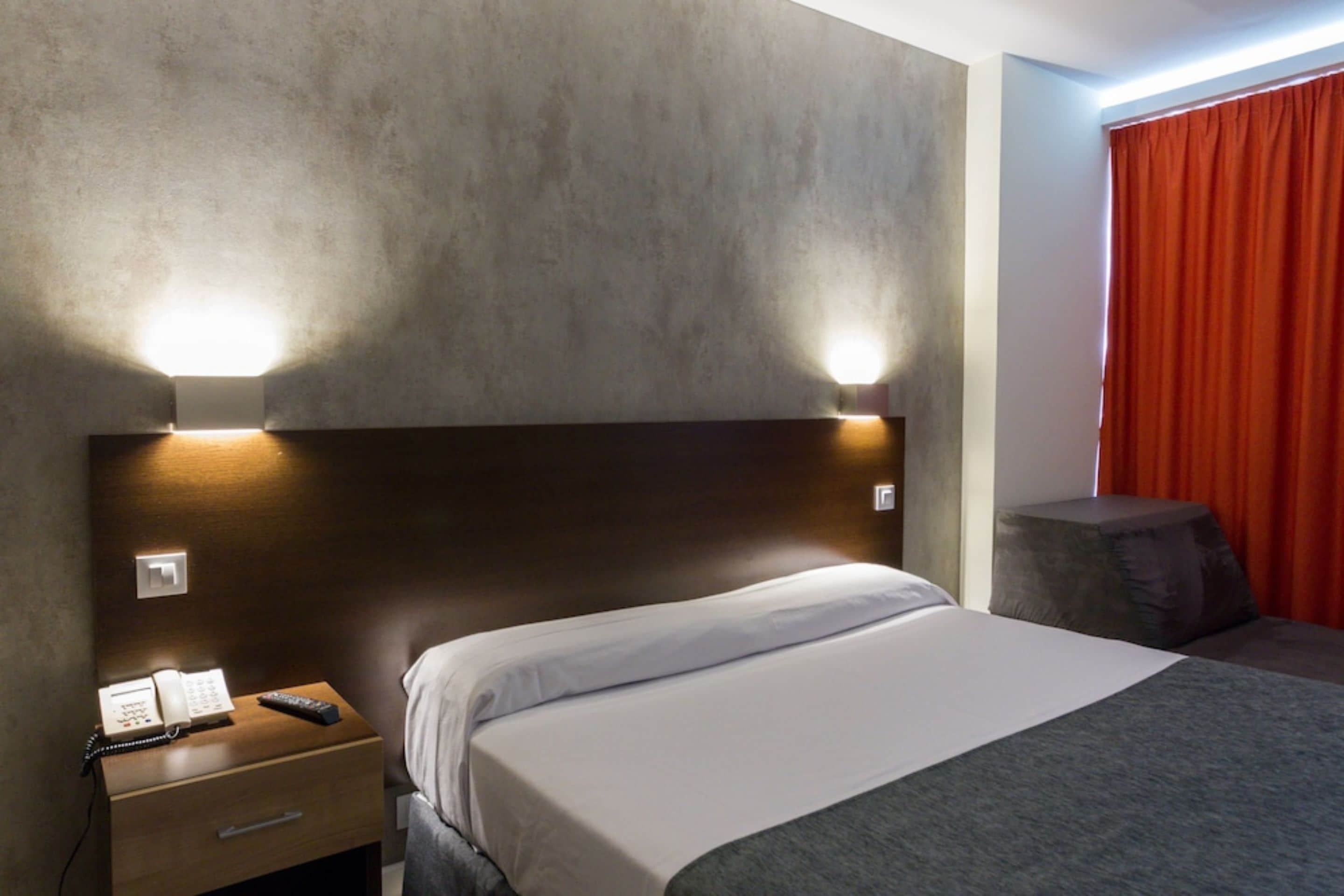 A hotel room with a bed, a nightstand with a phone and remote, and a red curtain.