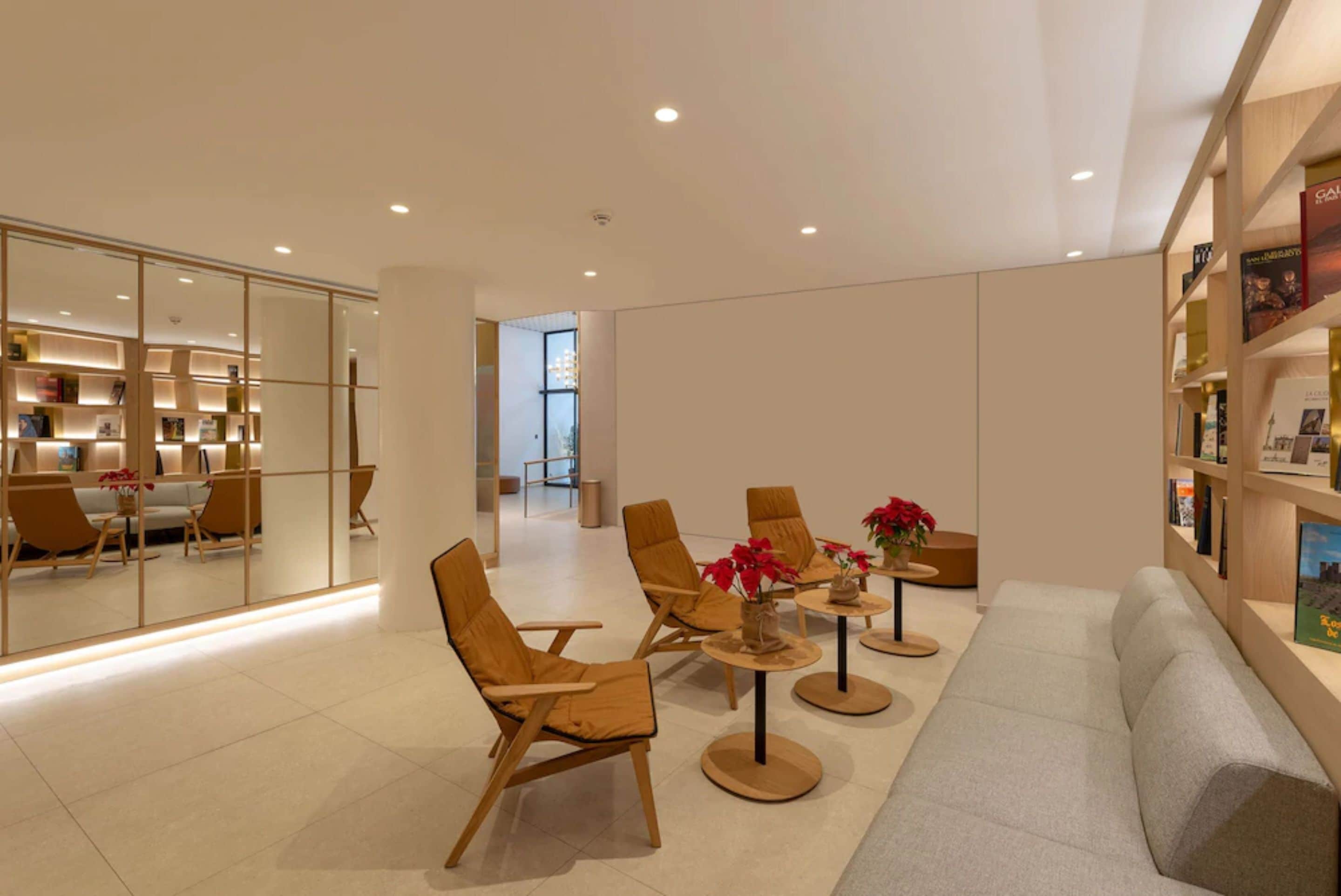 Un intérieur moderne avec un canapé, des fauteuils et des tables disposés pour créer un espace de détente. Des étagères sont alignées le long des murs et une cloison en verre se trouve à l’arrière-plan.