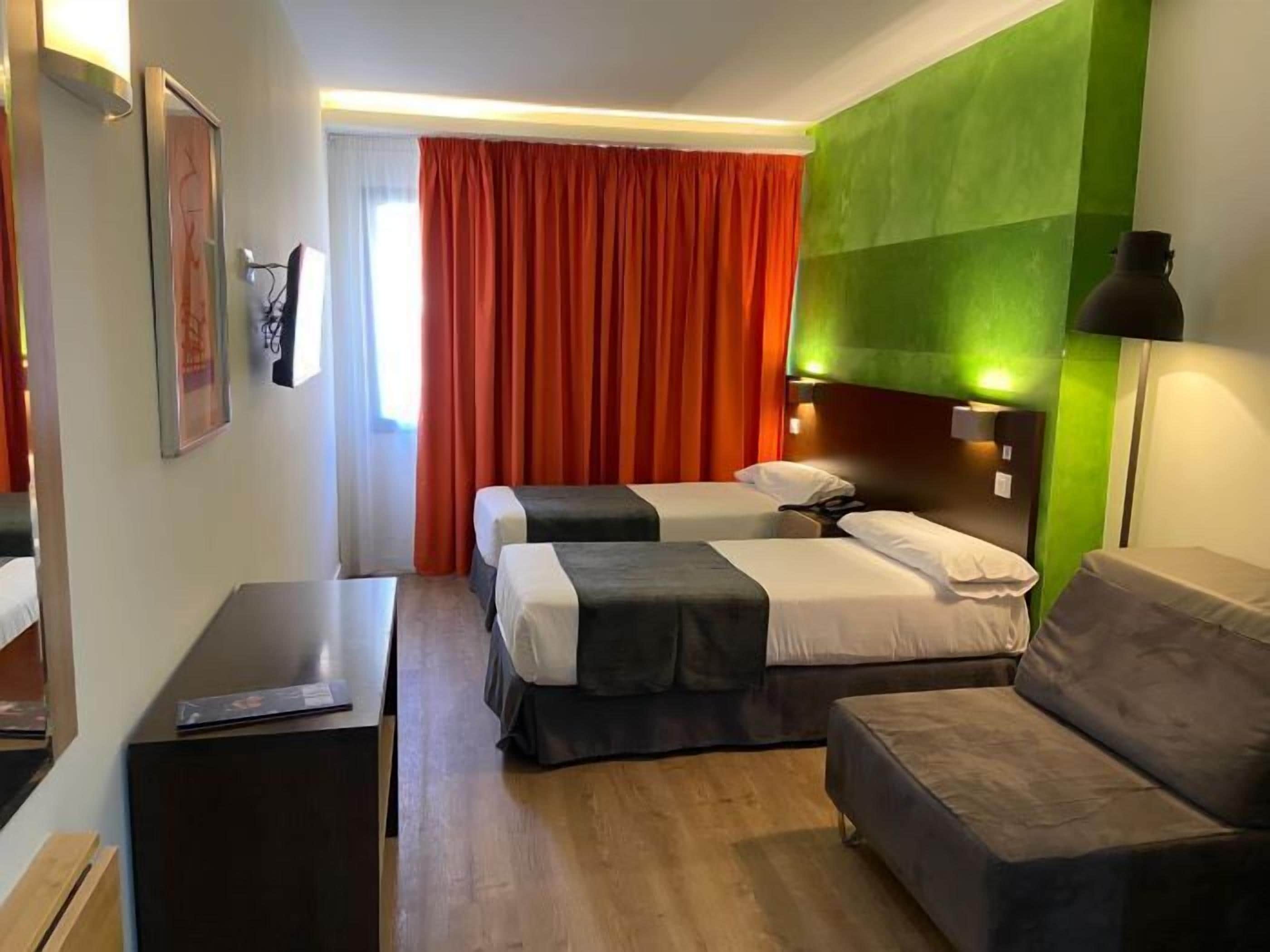 A hotel room with two beds, a sofa, a desk, and a green accent wall.