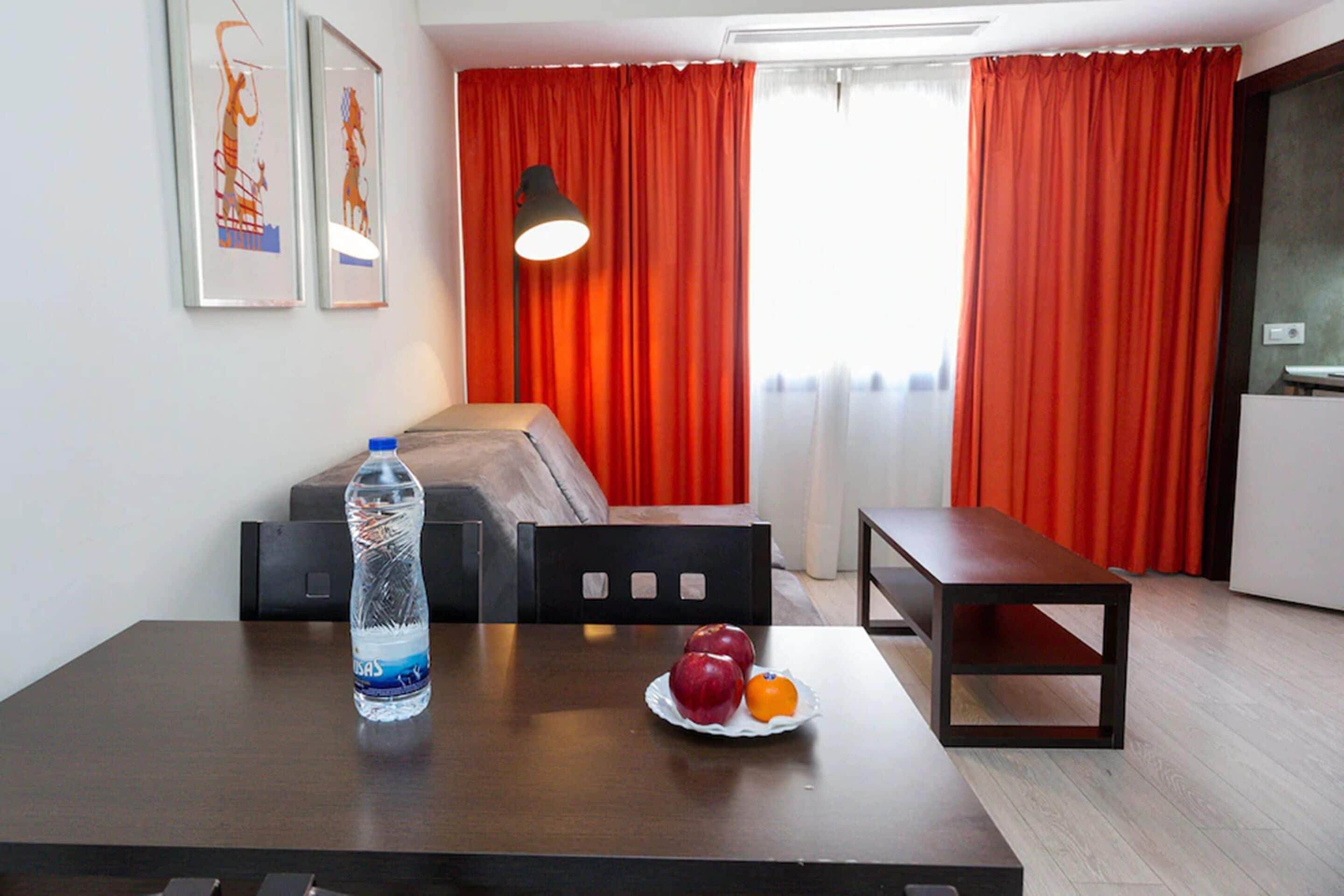 A hotel room with a bed, a table with a water bottle and fruit, a bench, and red curtains.