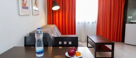 A hotel room with a bed, a table with a water bottle and fruit, a bench, and red curtains.