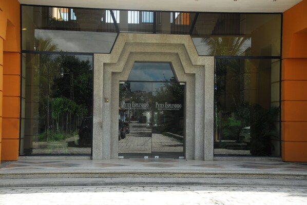 Entrada de hotel con fachada de vidrio, un arco de concreto y el nombre 'Peten Esplendido' exhibido en el vidrio.