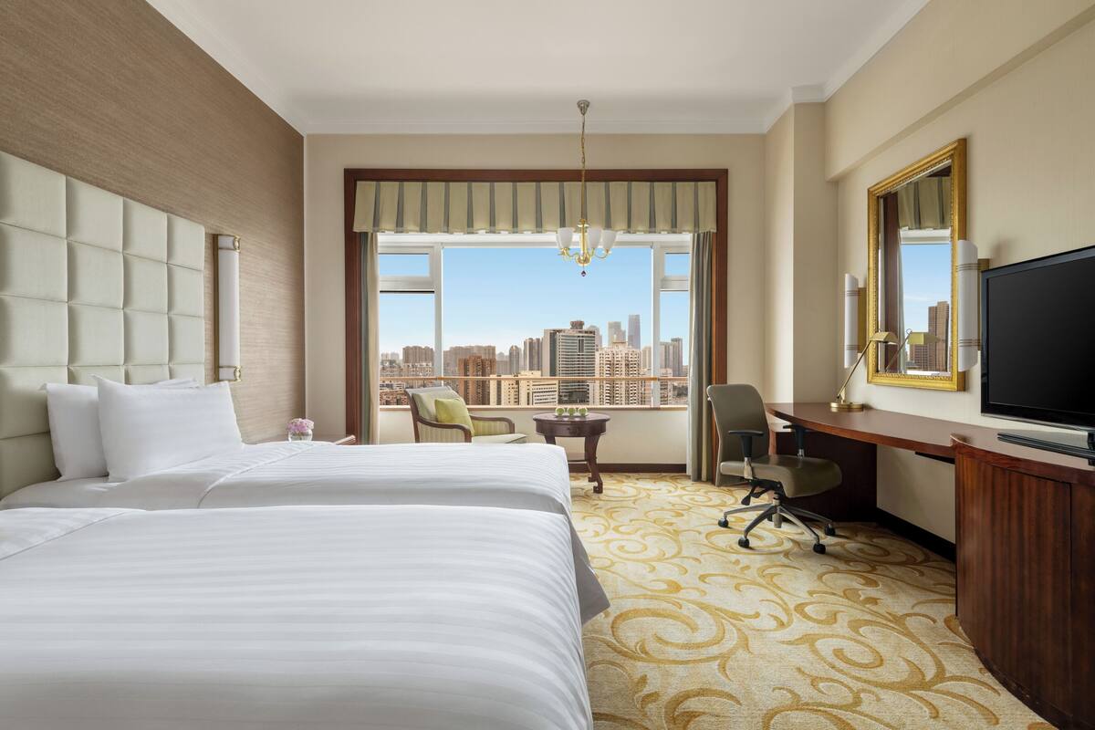 A hotel room with two beds, a desk, a chair, a television, and a large window offering a city view.