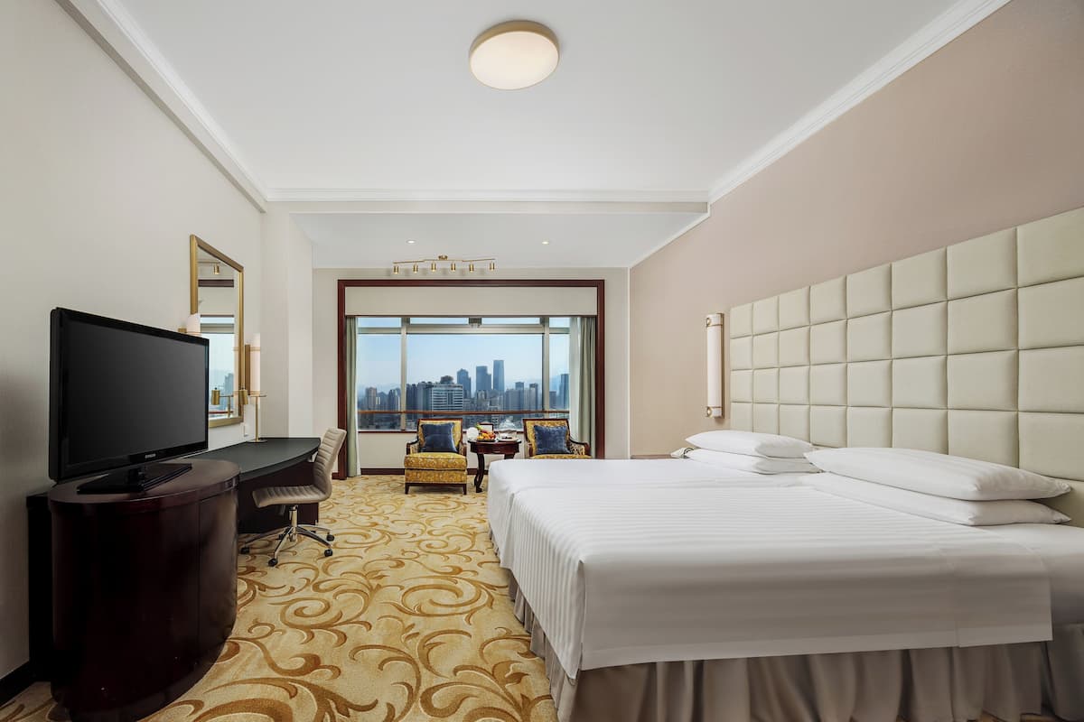 A hotel room with a large bed, a TV on a stand, a desk with a chair, and a view of the city through large windows.