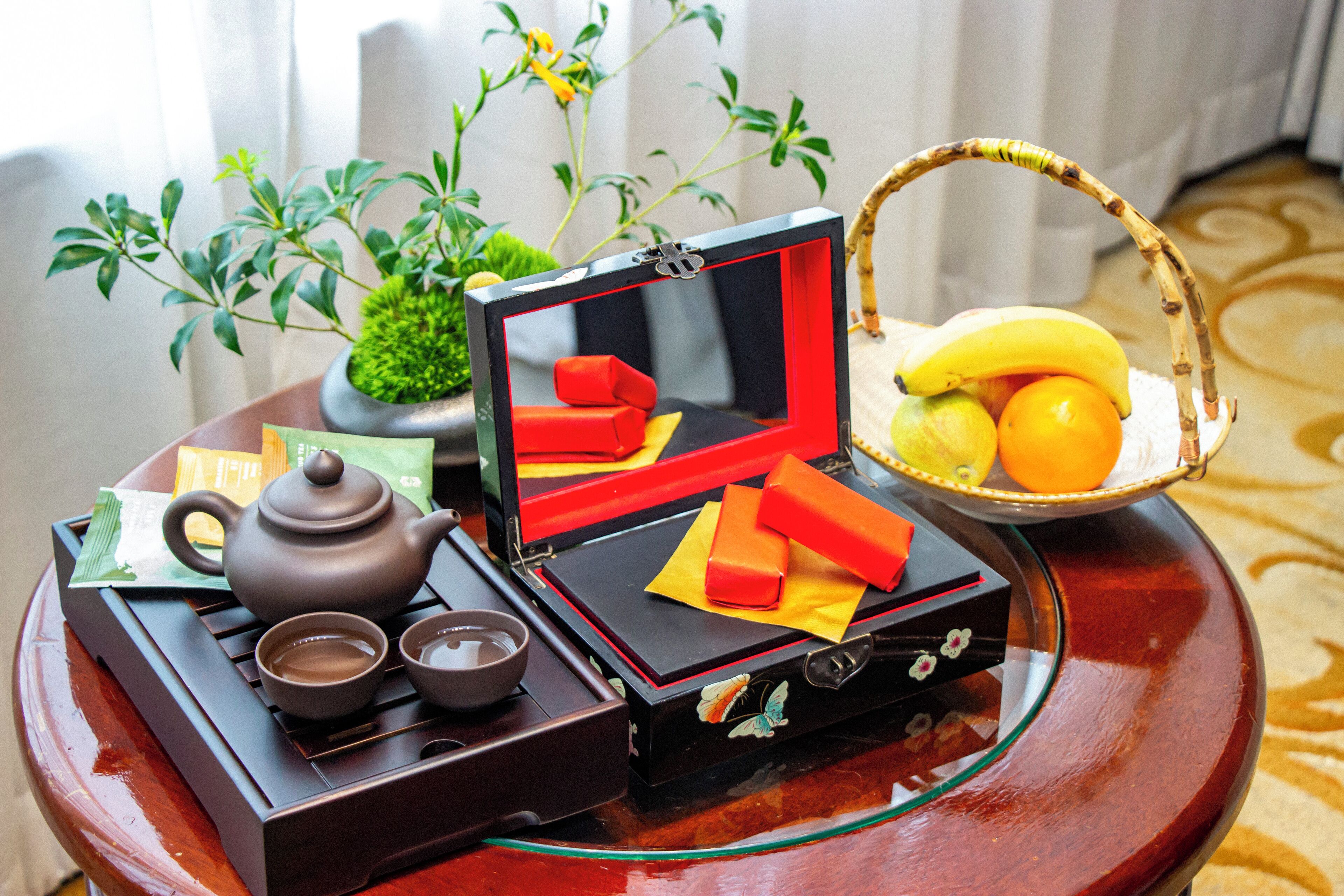 Een traditionele theeset met theepot, kopjes en een dienblad, een kleine potplant en een mand met fruit op een houten tafel.