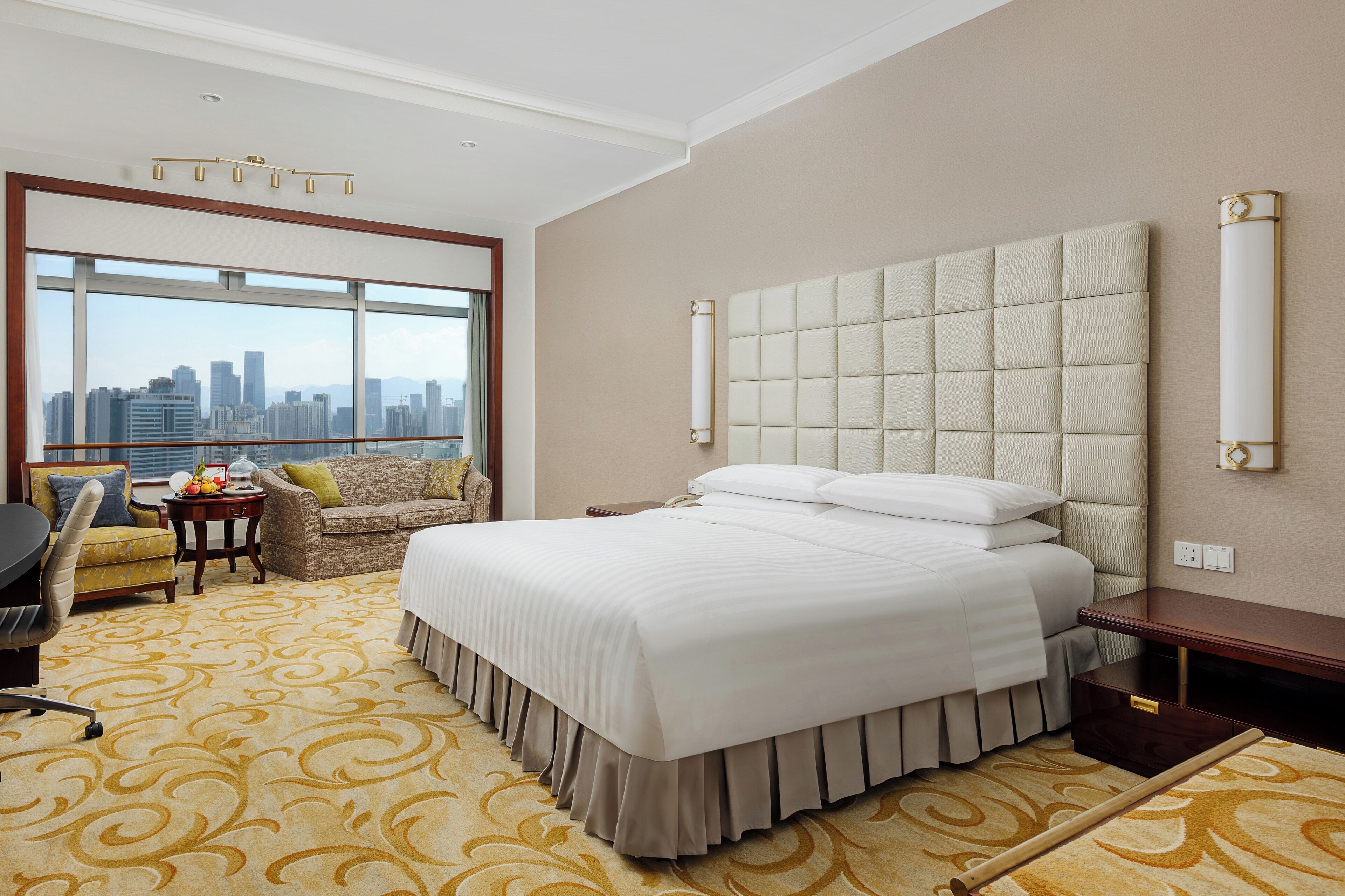 Une chambre d’hôtel dotée d’un grand lit, d’un canapé, d’un bureau et offrant une vue sur les toits de la ville grâce à de grandes fenêtres.