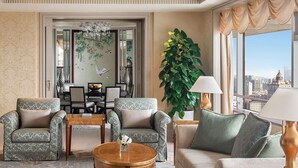 A spacious living room with a view of the city, featuring a chandelier, a large plant, and a dining area in the background.