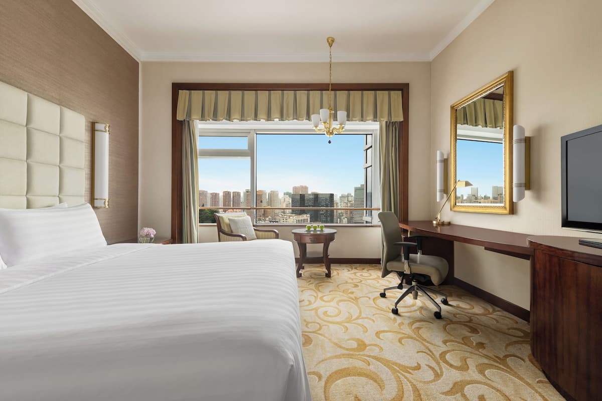 A hotel room with a large bed, a desk, a chair, a television, and a view of the cityscape.