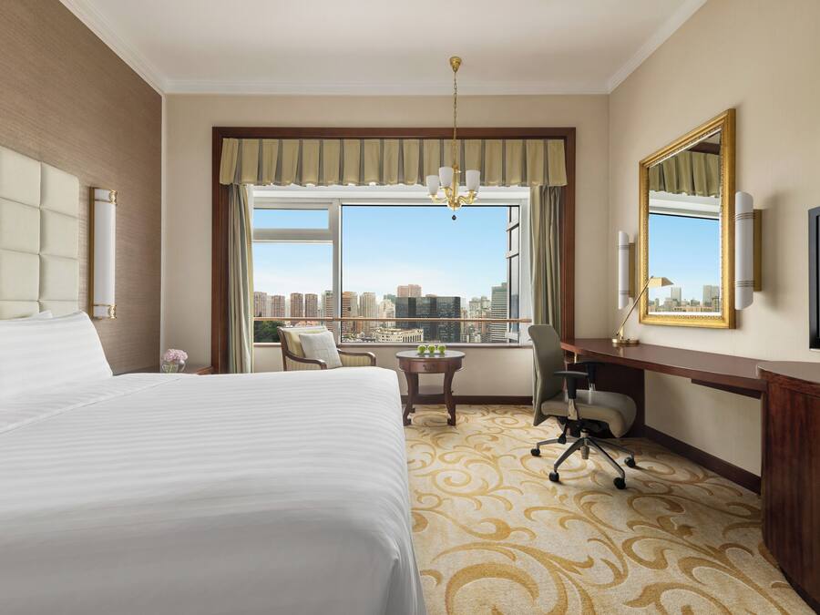 A hotel room with a large bed, a desk, a chair, a television, and a view of the cityscape.