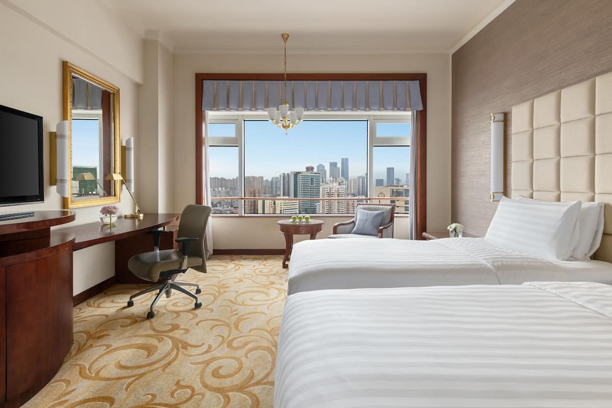 A hotel room with a large bed, a desk with a computer, a chair, and a view of a city skyline through a large window.