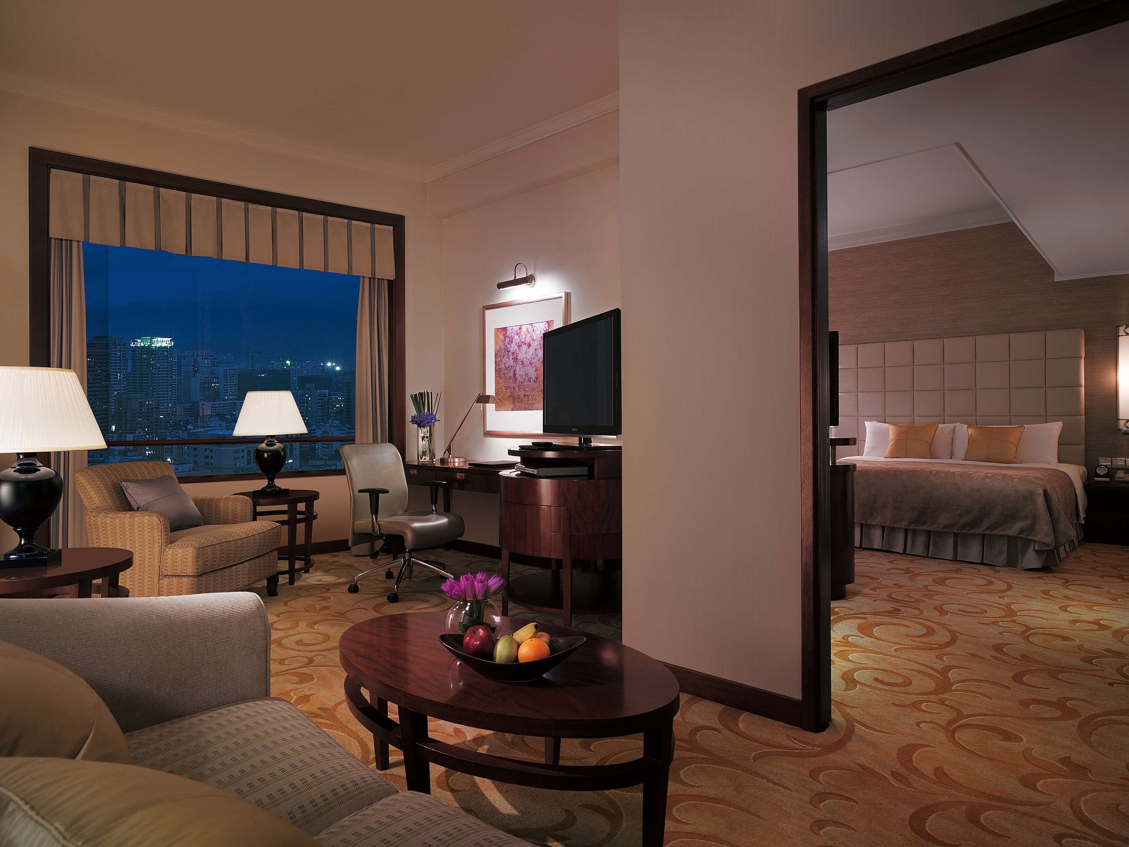 A hotel room with a city view, a desk with a computer, a TV, a bed, and a dining area with a table and chairs.