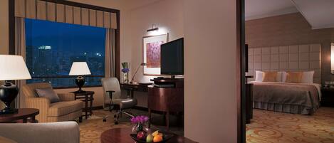 A hotel room with a city view, a desk with a computer, a TV, a bed, and a dining area with a table and chairs.