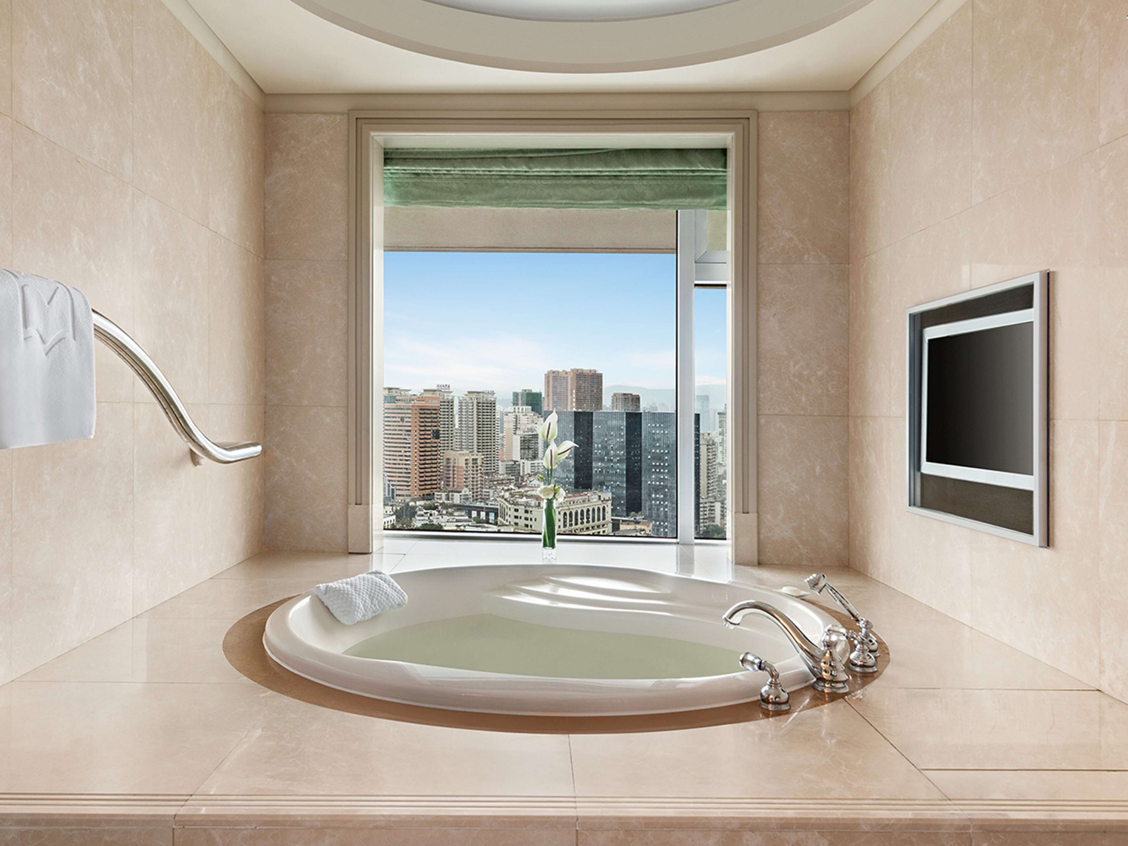 A modern bathroom with a large bathtub, a view of a cityscape, and a flat-screen TV mounted on the wall.