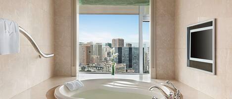 A modern bathroom with a large bathtub, a view of a cityscape, and a flat-screen TV mounted on the wall.