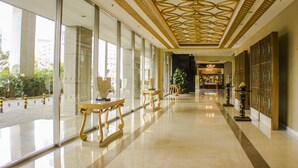 A spacious, well-lit corridor with large windows, elegant gold-framed furniture, and a decorative ceiling.