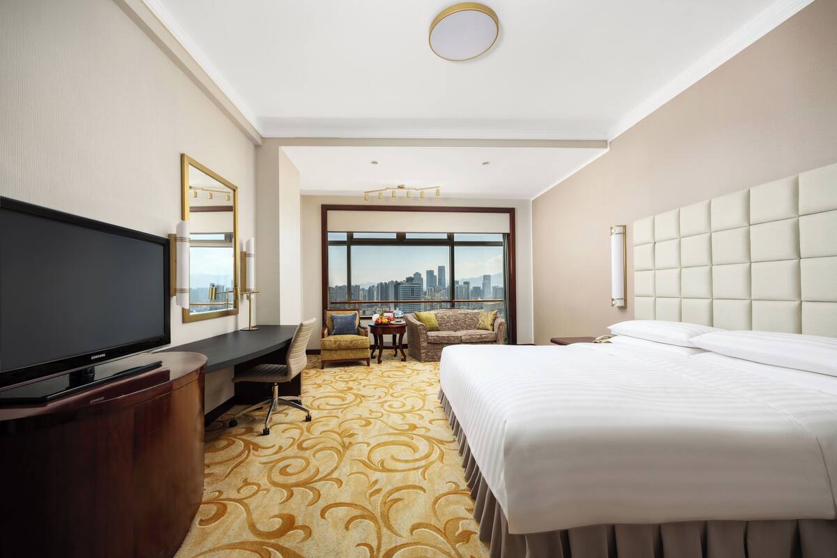 A hotel room with a large bed, a television, a desk, and a view of the city.