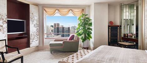 Ein geräumiges Hotelzimmer mit einem großen Bett, einem Fernseher, einer Sitzecke mit Stuhl und Sofa, einer großen Kübelpflanze und Blick auf die Stadt durch das Fenster.