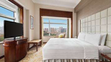 A hotel room with a large bed, a television on a wooden stand, a small table, and a view of the city through a large window.