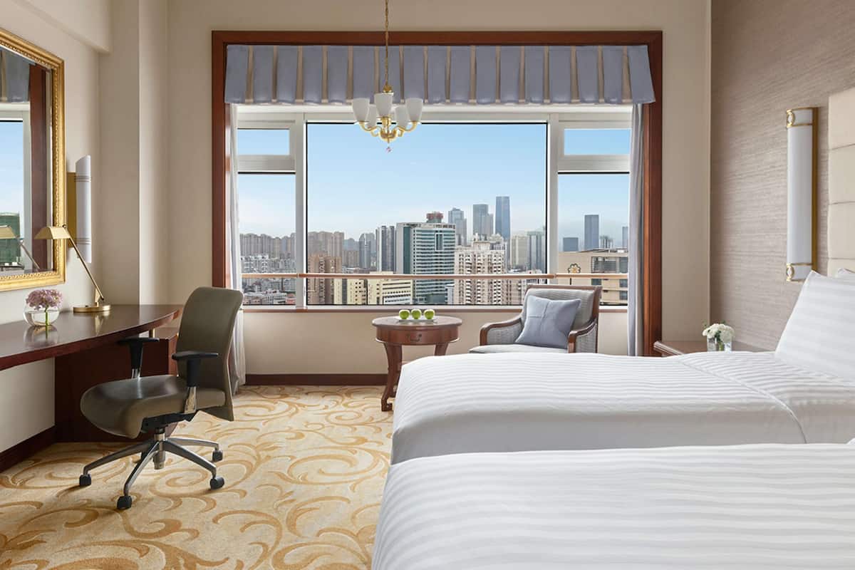 A hotel room with a large bed, a desk with a chair, a small table with a vase, and a view of a cityscape through large windows.