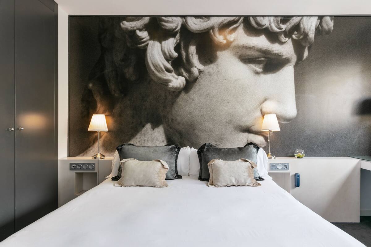 A modern hotel room with a large wall mural, a neatly made bed with pillows, bedside lamps, and a small table with a bowl of fruit.