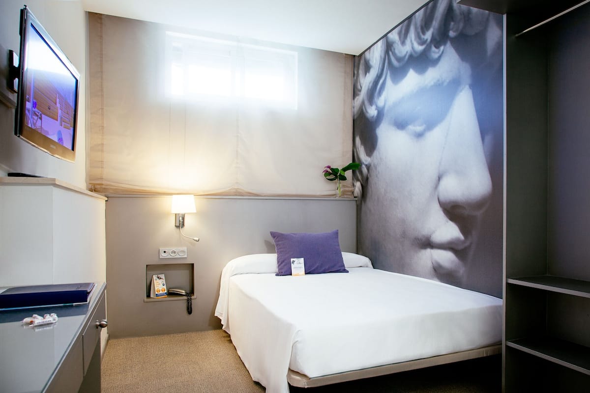 A hotel room with a bed, a TV mounted on the wall, a bedside table with a lamp, a small shelf with a phone and a brochure, and a large wall mural.