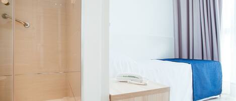 Une chambre d’hôtel avec une douche en verre, une table de chevet avec un téléphone et une télécommande, un lit avec une couverture bleue et un rideau.