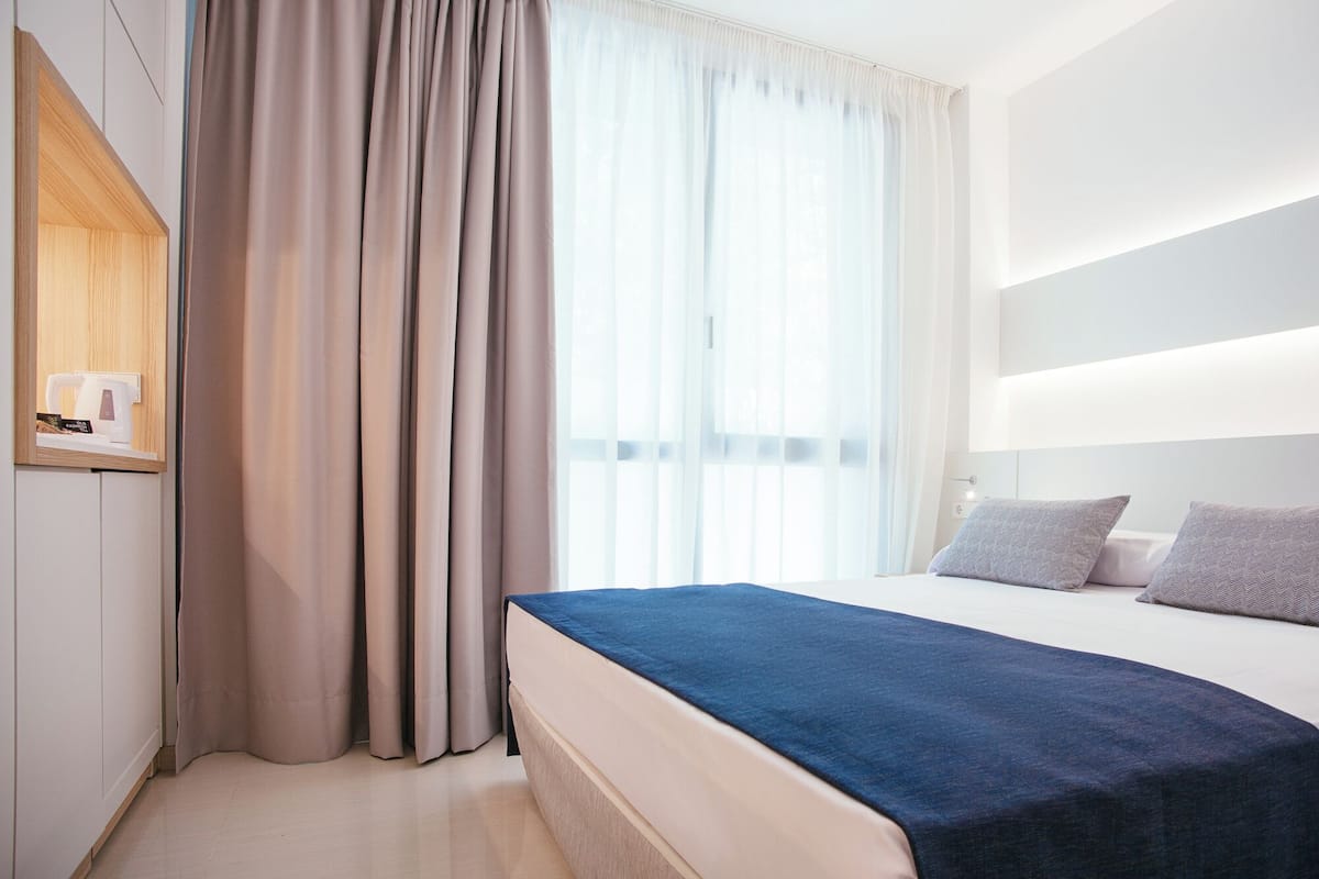 A modern hotel room with a bed, a nightstand, a mirror, and a window with curtains.