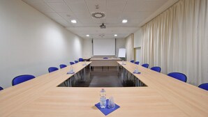 Een vergaderruimte met een lange tafel, blauwe stoelen, een whiteboard en een projectiescherm.