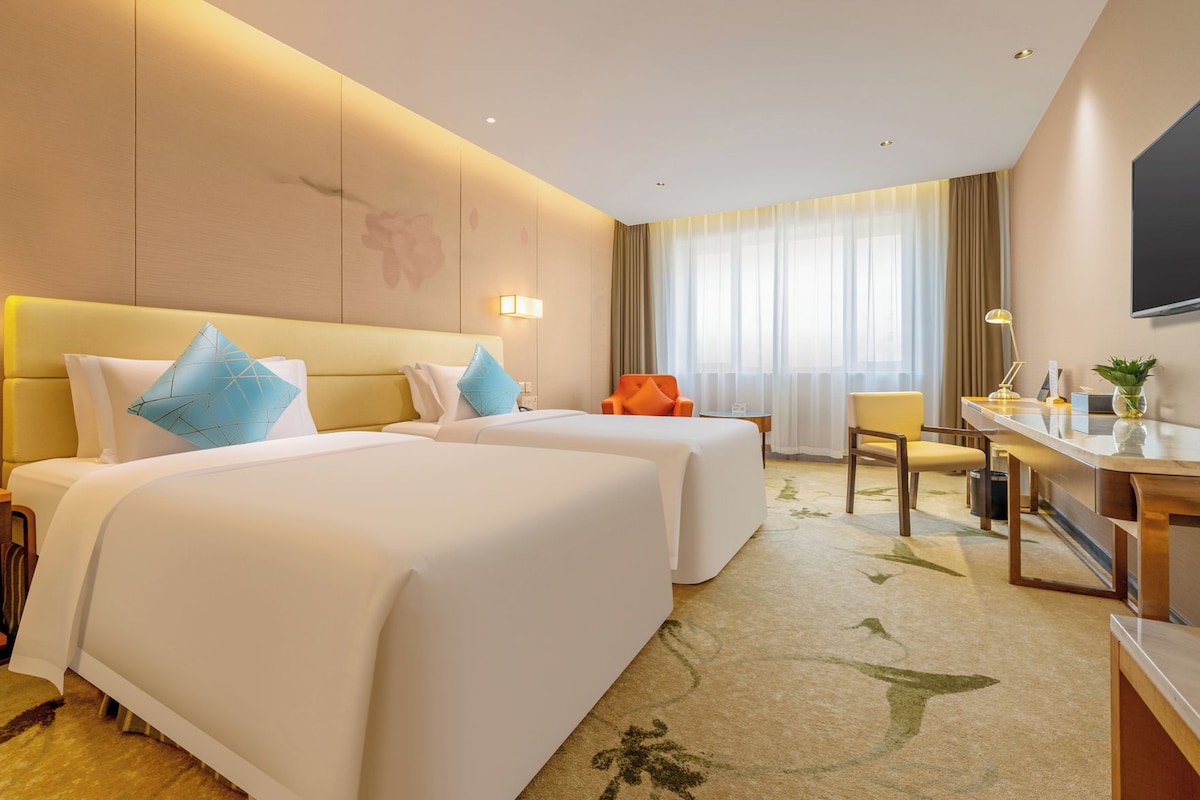 Une chambre d’hôtel avec deux lits, un bureau, une chaise, une télévision et une grande fenêtre avec des rideaux.