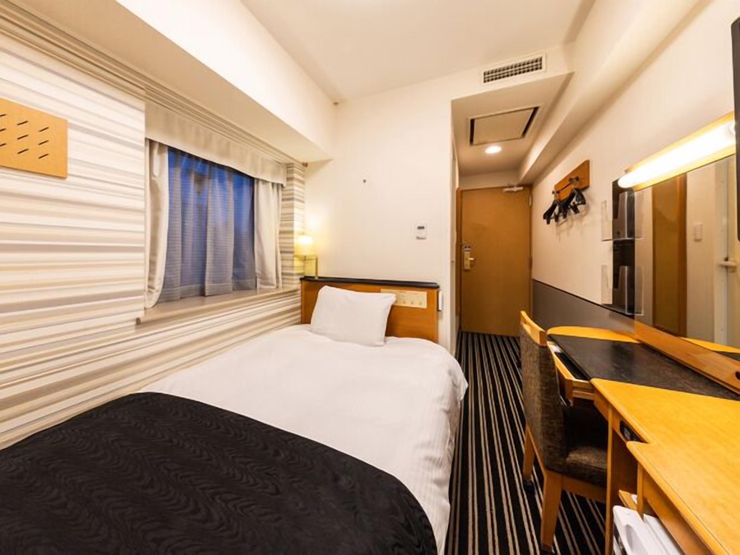 A hotel room with a bed, a desk, a mirror, and a window with curtains.