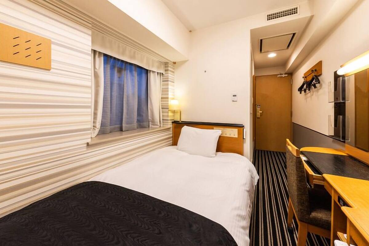 A hotel room with a bed, a desk, a mirror, and a window with curtains.