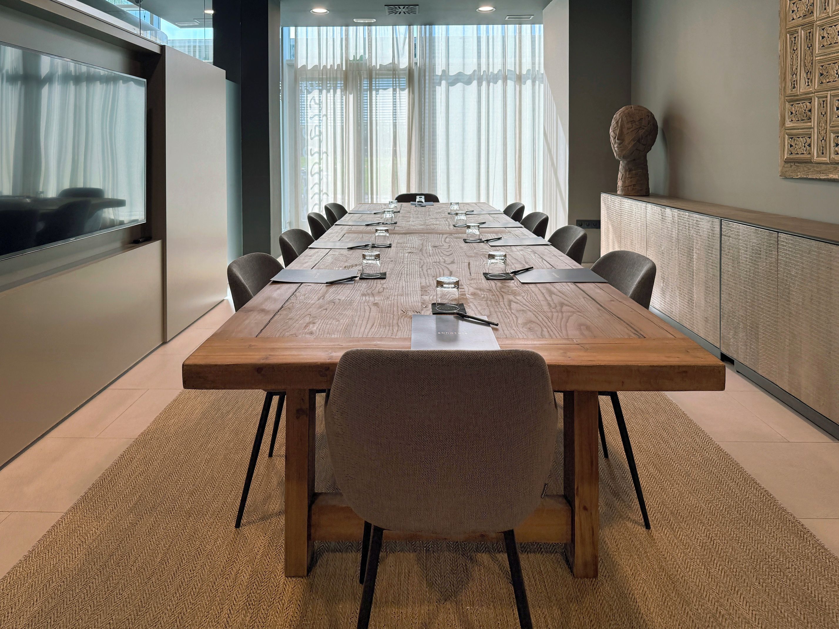 A conference room with a long wooden table, black chairs, and a large window with sheer curtains.