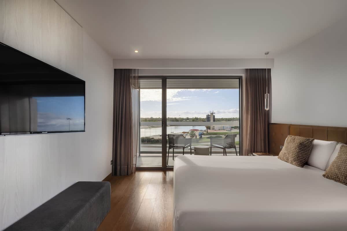 Quarto de hotel com cama, televisão na parede, varanda com vista para a cidade e área de jantar com cadeiras.