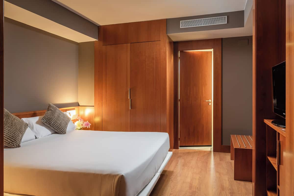 Quarto de hotel com cama, guarda-roupa de madeira, televisão e um banco.