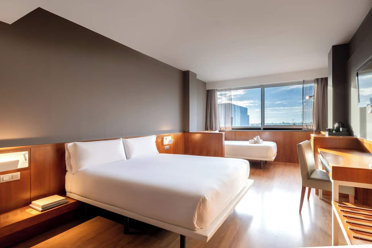 Quarto de hotel com cama, mesa de cabeceira, escrivaninha, cadeira e uma grande janela com vista para a cidade.