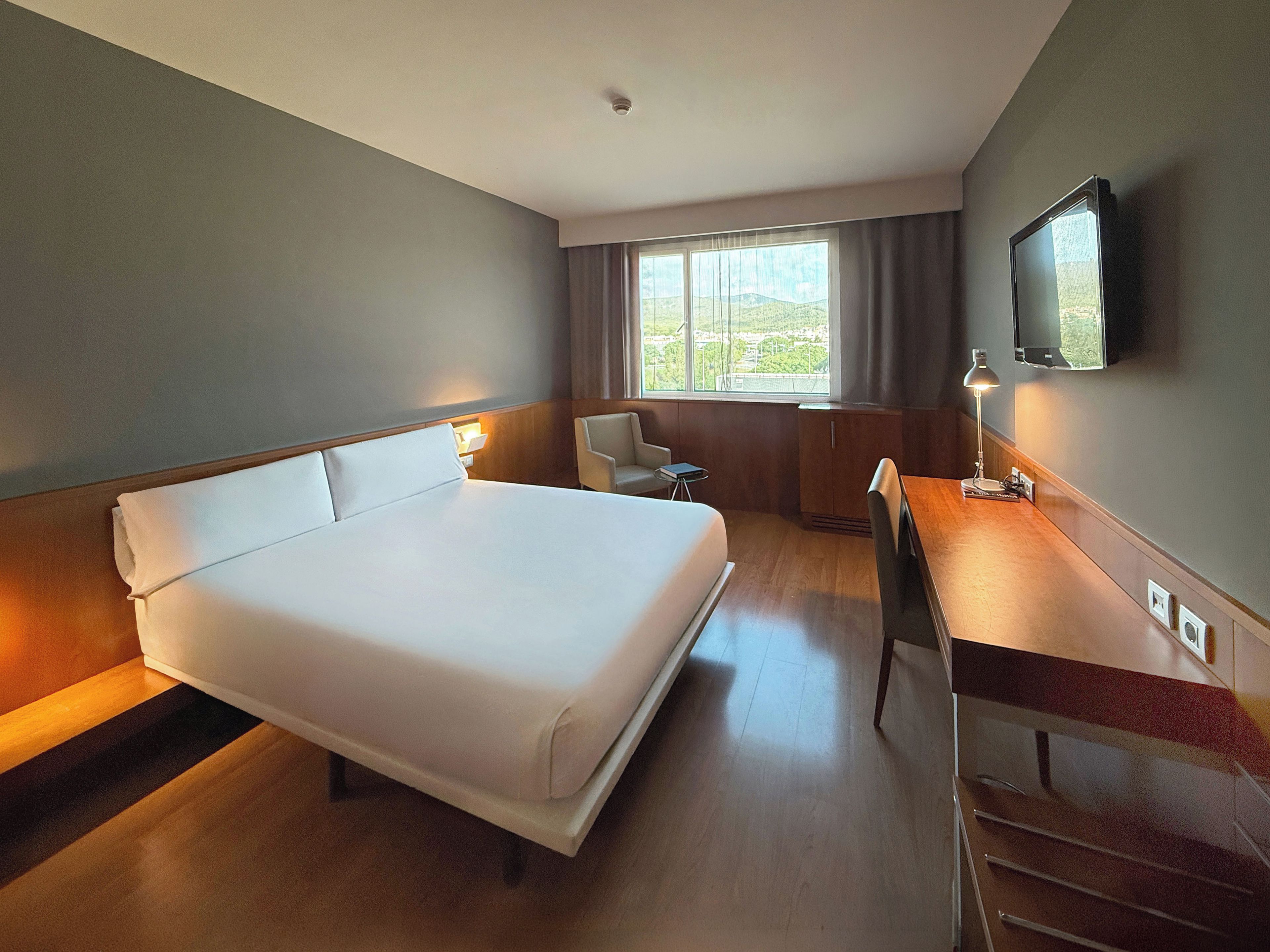 A hotel room with a large bed, a desk, a chair, a TV, and a window with curtains.
