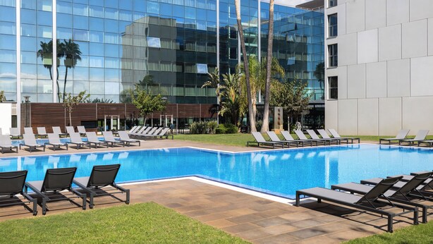 A modern building with a reflective glass facade, a swimming pool with lounge chairs, and palm trees. - Hotel SB BCN Events 4 Sup (Castelldefels)
