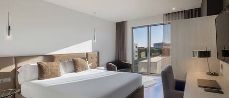 A modern hotel room with a large bed, a desk, a chair, a TV, and a view of the city.