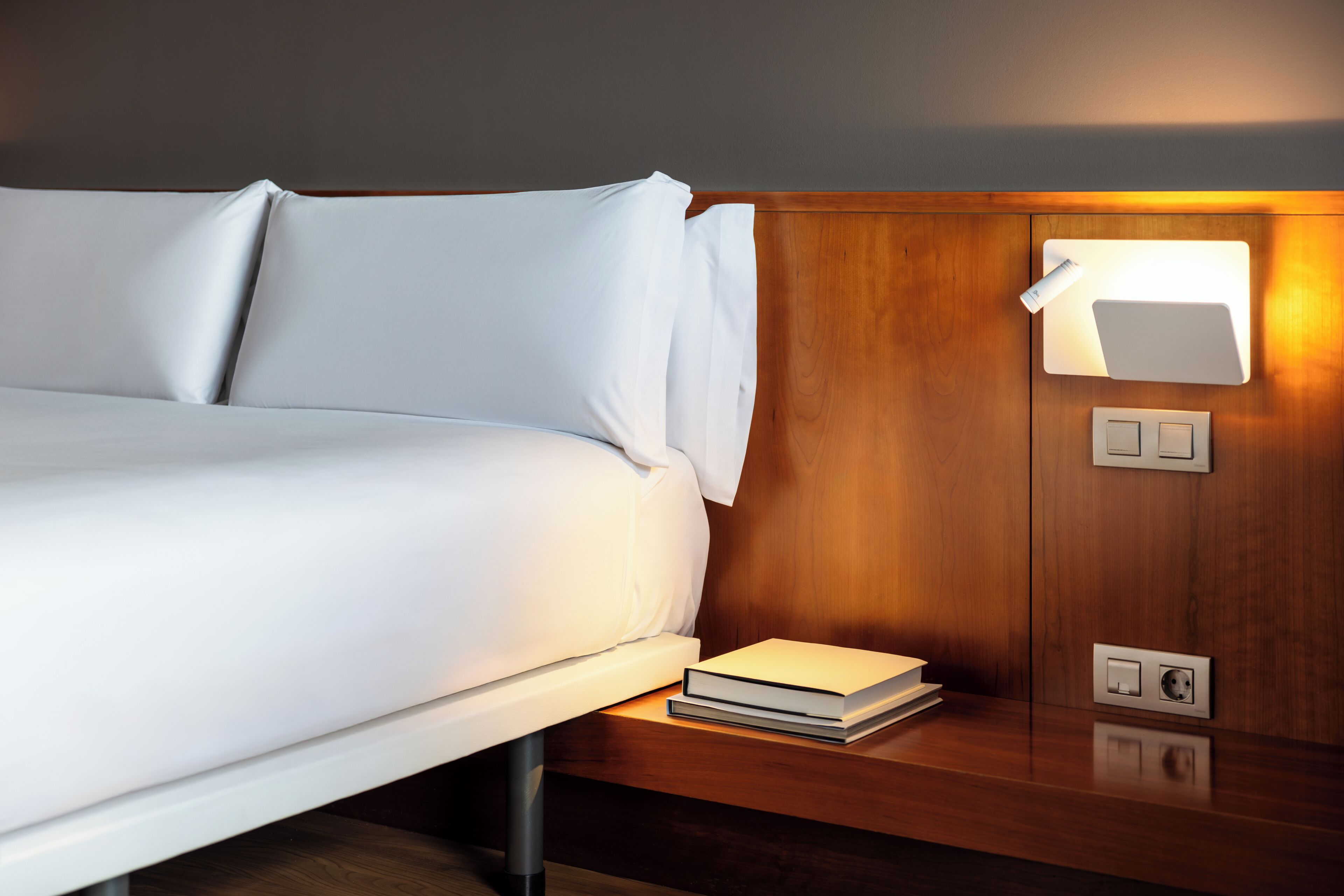 Una cama bien tendida con ropa de cama blanca, una mesita de noche con un libro, y una lámpara de pared.
