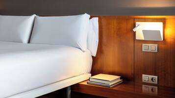 Una cama bien tendida con ropa de cama blanca, una mesita de noche con un libro, y una lámpara de pared.